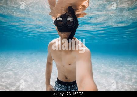 Der Freibeuter in der Tauchermaske macht Selfie unter Wasser über sandigem Boden in blauem Ozeanwasser. Aktivitätsurlaub im tropischen Meer. Stockfoto