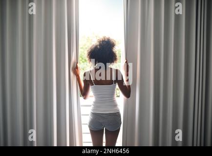 Raus aus den Federn. Rückblick auf eine junge Frau, die die Vorhänge in ihrem Schlafzimmer öffnete. Stockfoto