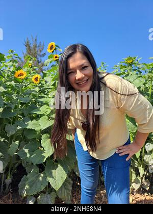 Lateinische Erwachsene Frau mit Sonnenbrille geht durch ein Feld voller Sonnenblumen und vergisst ihre Probleme voller Glück in Fülle, mit Ruhe, Entspannung Stockfoto