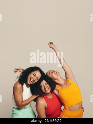 Drei junge Frauen lachen fröhlich, während sie in Sportkleidung in einem Studio zusammenstehen. Eine Gruppe weiblicher Freunde, die ihren fit, gesund feiern Stockfoto