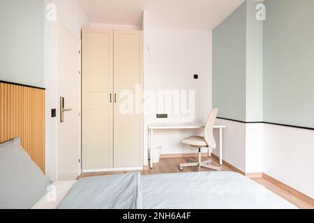 Ein heller Raum mit Wänden in sanften Minztönen, ein Bett für zwei Personen, ein Arbeitsbereich mit Tisch und Stuhl, ein Kleiderschrank mit beigen Holztüren. Tür Stockfoto