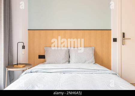 Nahaufnahme des Bettes mit Kopfteil an der Wand mit Holzdekor. Das Bett ist in Pastellgrau gehalten. Großes Fenster mit dunkelgrauen Vorhängen. Nachttisch mit Stockfoto