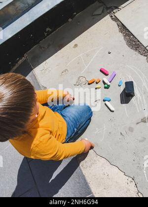 Kleinkind in Jeans zieht an sonnigen Tagen mit Buntstiften auf den Asphalt. Das Kind hält Buntstifte in der Hand. Die Hände und Kleidungsstücke des Kindes sind mit bunten Flecken bedeckt. Stockfoto