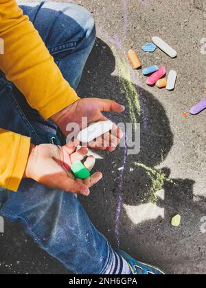 Kleinkind in Jeans zieht an sonnigen Tagen mit Buntstiften auf den Asphalt. Das Kind hält farbige Buntstifte in der Hand. Kinderkleidung ist mit bunten Flecken bedeckt Stockfoto