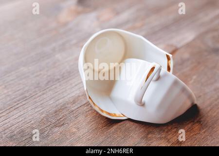 Zerbrochene weiße Tasse auf Holzhintergrund. Beschädigte Tasse mit goldener Dekoration. Stockfoto