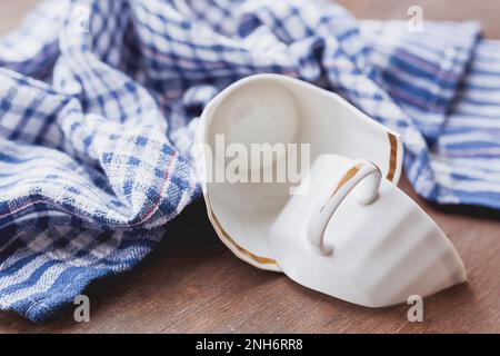 Zerbrochene weiße Tasse auf Holzhintergrund mit blauem Karottentuch. Beschädigte Tasse mit goldener Dekoration. Stockfoto