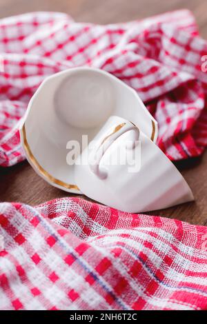 Zerbrochene weiße Tasse auf Holzhintergrund mit rotem kariertem Handtuch. Beschädigte Tasse mit goldener Dekoration. Stockfoto