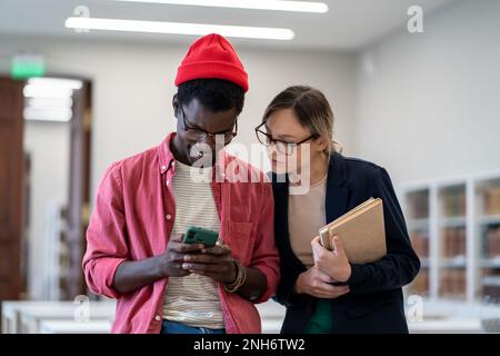Ein paar Studenten gemischter Rassen schauen auf ein Smartphone und schauen sich im Break Study ein lustiges virales Videonetzwerk an Stockfoto