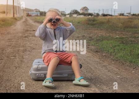 Das Kind schaut durch das Fernglas und sitzt auf einem Koffer vor dem Hintergrund der Straße. Ein Reisender, der im Urlaub spielt. Hochwertiges Foto Stockfoto