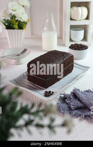 Schokoladenkuchen mit gedämpftem Schwamm vor dem Hintergrund einer Flasche Milch und Blumen in einer Vase Stockfoto