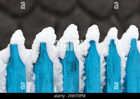 Der schneebedeckte alte Holzzaun ist blau, vor dem Hintergrund des grauen Fundaments des Hauses. Retro-Style bei Schneewetter Stockfoto