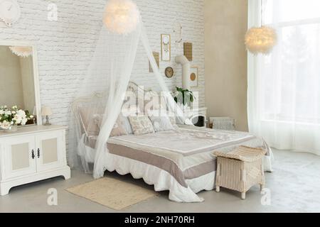 Komfortables, großes, einfaches Schlafzimmer mit hellen Möbeln, Himmelbett, Federlampen, kleinem Kamin auf Holz, gemütlichem, modernem Platz zum Ausruhen und Stockfoto
