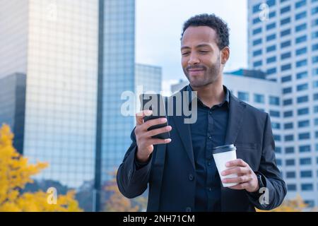 Erfolgreicher schwarzer Geschäftsmann im Anzug, der mit Smartphone und Kaffeetasse im Finanzviertel lächelt Stockfoto