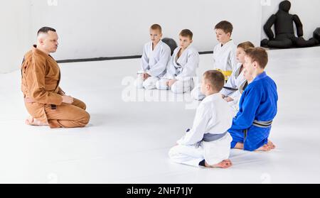 Eine Gruppe kleiner Jungs, Kinder im Kimono, die auf dem Boden um die Kutsche sitzen. Kinder trainieren im Innenbereich. Judo, Jiu-Jitsu-Training Stockfoto