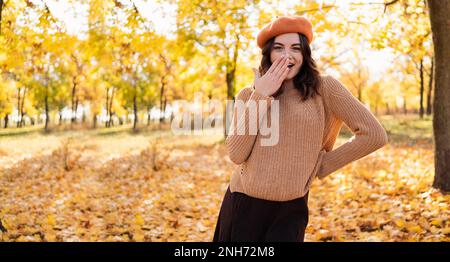 Porträt einer fröhlichen jungen Frau im Park im Herbst. Fröhliches Mädchen mit Pullover und orangefarbener Baskenmütze, die draußen freundlich lächelt. Hintergrund Herbst. Kopieren Sie Spac Stockfoto