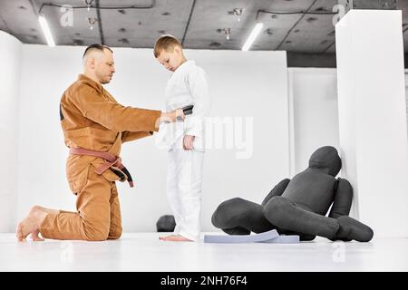 Mann, Judo, Jiu-Jitsu-Trainer, der dem Jungen hilft, der den Gürtel an Kimono bindet. Trainingsaktivität, lernen zu kämpfen Stockfoto