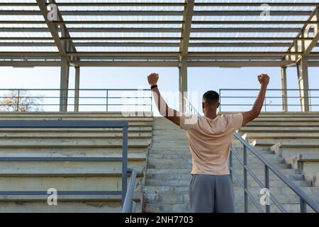 Ein lateinamerikanischer Sportler, der mit seinen Trainingsergebnissen, die erfolgreiche sportliche Leistung feiern, zufrieden ist. Der Läufer steht mit dem Rücken neben der Treppe und hält die Hände nach oben. Stockfoto