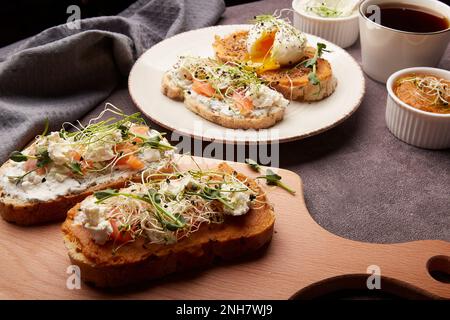 Ästhetisches, gesundes Frühstück - Sandwiches mit Lachs, pochiertem Ei, Zucchini und Frischkäse, Luzerne, Erbsen, Knoblauchcreme, Kaffee Stockfoto