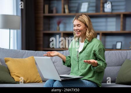 Fröhliche und glückliche Erwachsene Frau allein zu Hause, die auf dem Sofa im Wohnzimmer sitzt und den Laptop für Videoanrufe nutzt, die Hausfrau lächelt und sich mit Freunden aus der Ferne unterhält. Stockfoto