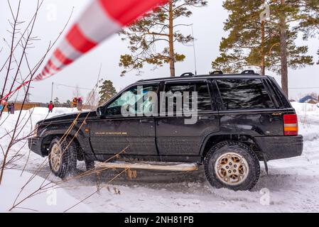 Offroad-SUV „Jeep Grand Cherokee“ in schwarzer Geländewagen fährt im Winter auf Schnee im Wald zwischen Kiefern und gestreiftem Klebeband mit einer Fahrerin. Stockfoto