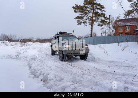 Offroad-SUV „Jeep Grand Cherokee“ in schwarzer Geländewagen fährt im Winter auf dem Schnee im Wald in der Nähe eines Familienhauses und eines Zauns mit einer Fahrerin. Stockfoto