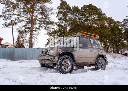 Geländewagen „Suzuki Escudo“ 4x4 mit Geländetraining steht auf einer verschneiten Straße im Wald vor dem Hintergrund von Bäumen und einem Zaun. Stockfoto