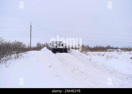 Der russische SUV „UAZ Patriot“ fährt im Winter schnell auf einer verschneiten Straße auf dem Feld vor dem Hintergrund des Waldes, der am Schneesturmrad klebt. Stockfoto