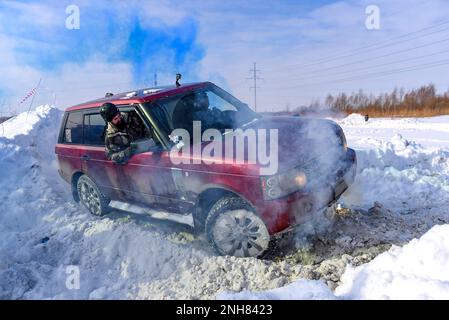 Ein „Range Rover 4x4“-SUV fährt aktiv in blauem Rauch auf einem verschneiten Berg, und ein Mann mit Schutzhelm beobachtet draußen die Straße. Stockfoto