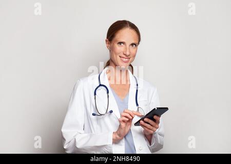 Selbstbewusst lächelnde moderne mittelErwachsene Frau Arzt Arzt Praktizierende mit Smartphone, weibliches Porträt Stockfoto