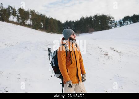 Bärtiger Mann mit einem Rucksack in Sonnenbrille und warmer Kleidung vor dem Hintergrund der Winterberge und des Waldes. Das Konzept der Wanderung und der aktiven Lif Stockfoto