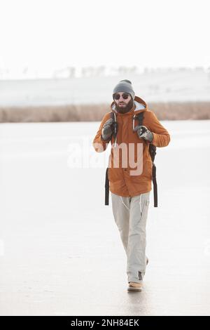 Ein bärtiger Mann mit einem Rucksack in warmer Winterkleidung läuft im Winter auf einem gefrorenen See. Das Konzept der Wanderung und des aktiven Lebensstils. Stockfoto