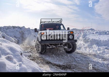 Russischer Offroad-Geländewagen „Lada Niva 4x4“ mit schnellem Schneesturm, der im Winter mit einem fröhlichen Fahrer in einem Hut mit Ohrenschützern durch den Schnee fährt. Stockfoto
