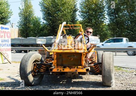 Ein lächelnder Mann mit dunkler Brille sitzt in einem kleinen provisorischen Buggy, der am Straßenrand fährt Stockfoto