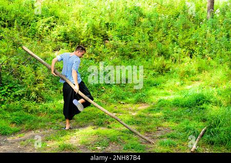 Der Mann in der breiten Hose, der Yoga macht, dreht den großen Stock im Wald auf dem Gras. Stockfoto