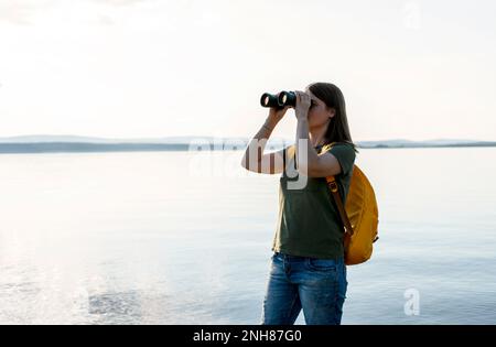 Junge Frau mit gelbem Rucksack, die durch ein Fernglas Vögel am See oder Fluss ansieht Vogelbeobachtung, Zoologie, Ökologie Forschung in der Natur, Beobachtung o Stockfoto