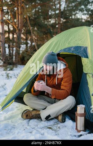 Ein junger bärtiger Mann ruht in den Winterbergen in der Nähe eines Zelts. Ein Reisender mit einem Bart in einer Mütze und einer warmen Jacke wärmt sich auf, indem er heißen Tee oder trinkt Stockfoto