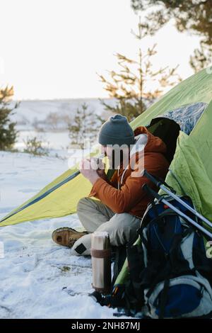 Ein junger bärtiger Mann ruht in den Winterbergen in der Nähe eines Zelts. Ein Reisender mit einem Bart in einer Mütze und einer warmen Jacke wärmt sich auf, indem er heißen Tee oder trinkt Stockfoto