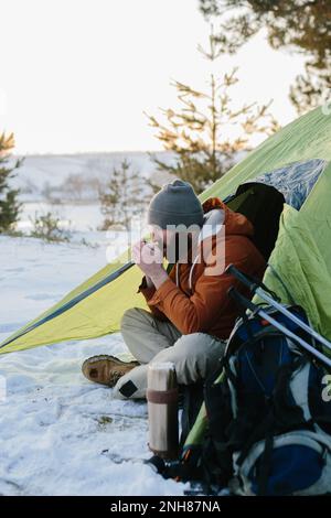 Ein junger bärtiger Mann ruht in den Winterbergen in der Nähe eines Zelts. Ein Reisender mit einem Bart in einer Mütze und einer warmen Jacke wärmt sich auf, indem er heißen Tee oder trinkt Stockfoto