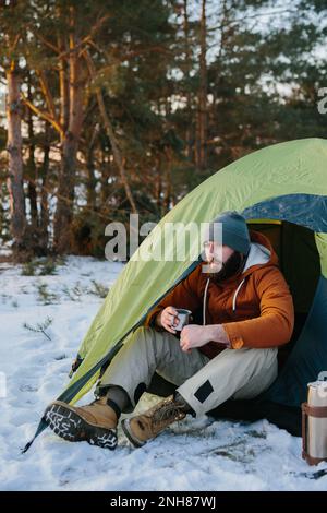 Ein junger bärtiger Mann ruht in den Winterbergen in der Nähe eines Zelts. Ein Reisender mit einem Bart in einer Mütze und einer warmen Jacke wärmt sich auf, indem er heißen Tee oder trinkt Stockfoto