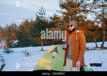 Ein Tourist, der mit einer Tasse Tee oder Kaffee am Zelt steht, genießt den Sonnenuntergang im Winterwald. Stockfoto