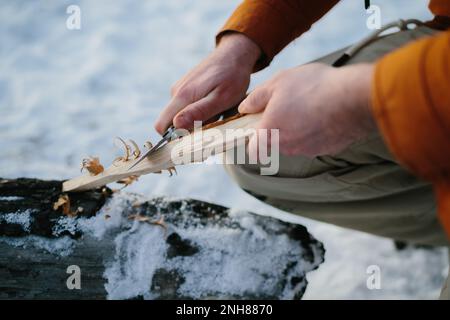 Ein Mann bereitet Holzwolle mit einem Messer zu, um bei Sonnenuntergang in einem Winterwald ein Feuer zu machen. Winterüberlebenskonzept. Stockfoto