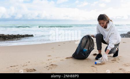 Junge Freiwillige, die Flaschen in Plastiktüte pflücken, schwarz, um morgens den Strand zu putzen, Panorama, freien Platz Stockfoto