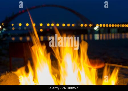 Die helle Flamme eines Lagerfeuers mit Funken an einem Sandstrand auf der Rückseite eines eisernen Grills und einer bogenförmigen Brücke in Novosibirsk mit bunten Lichtern Stockfoto