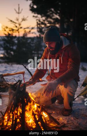 Ein Mann sitzt neben einem Lagerfeuer und wärmt sich bei Sonnenuntergang am Feuer in einem Winterwald. Stockfoto