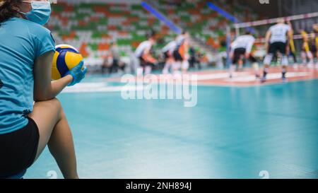 Mädchen mit Gesichtsmaske und Voleyball in den Händen im Hintergrund-Volleyballspiel. Stockfoto