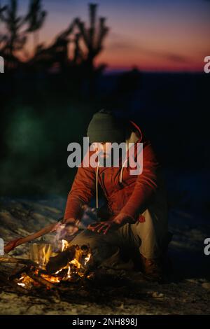 Ein Mann sitzt neben einem Lagerfeuer und wärmt sich bei Sonnenuntergang am Feuer in einem Winterwald. Stockfoto