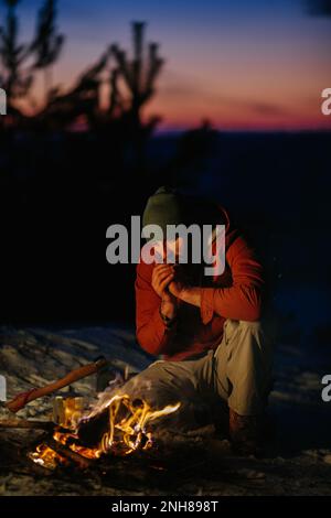 Ein Mann sitzt neben einem Lagerfeuer und wärmt sich bei Sonnenuntergang am Feuer in einem Winterwald. Stockfoto