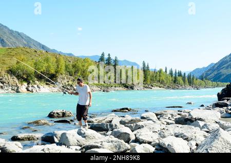 Männlicher Angler in aufgerollten Hosen, weißem T-Shirt und Mütze zum Angeln, warm und hell, schneller türkisfarbener Bergfluss Katun im Altai auf dem steinigen Stockfoto