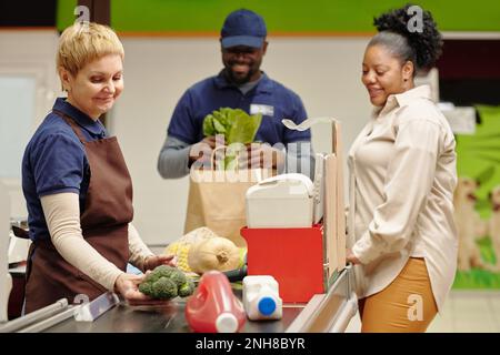 Glückliche, reife Verkäuferin steht an der Kasse im Supermarkt und bedient junge afroamerikanische Konsumentinnen Stockfoto