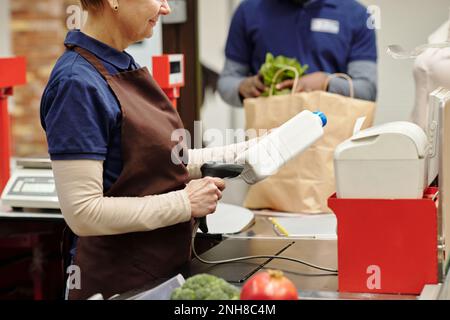 Seitenansicht des Kassierers, der den Barcode auf einer weißen Kunststoffflasche scannt, während er am Arbeitsplatz steht und einen der Kunden bedient Stockfoto